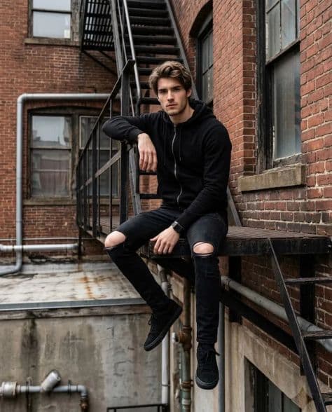 A model wearing layered urban streetwear posing against an old weathered red-brick factory wall.