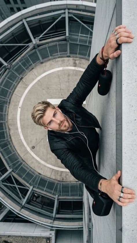 A man in tactical streetwear leaning on a steel industrial rooftop railing overlooking train tracks.