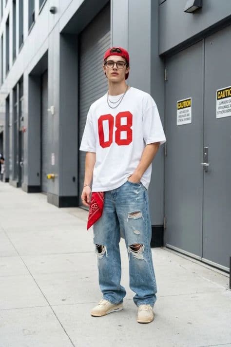 A young man in relaxed denim and jacket pausing naturally along a wide city sidewalk in an industrial district.
