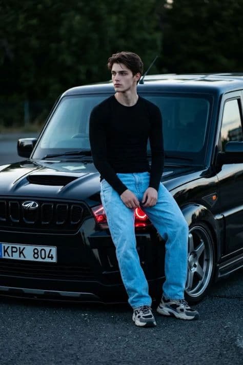 A person sitting on the front hood edge of an aggressive customized black Scorpio S11 SUV parked on a city street.