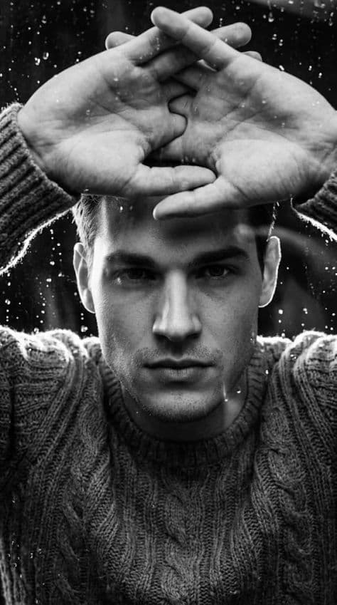 Black and white low-angle portrait of a man framed by interlocked hands behind a rain-streaked glass window