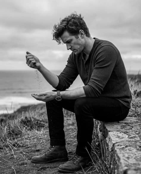 Black and white portrait of a man in a wool sweater seated on a coastal stone ledge