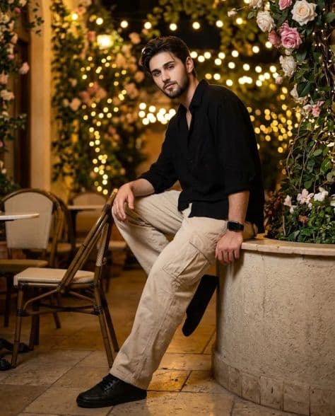 A casually dressed man seated in a garden café courtyard surrounded by glowing fairy lights and greenery.
