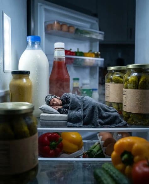 A tiny man sleeping on a miniature bed on a refrigerator shelf surrounded by everyday groceries.