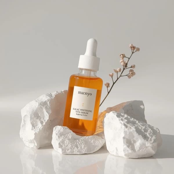 Minimal luxury skincare serum bottle labeled Ma:nyo Galac Whitening Vita Serum placed between white stones with warm sunlight and soft beige studio background.