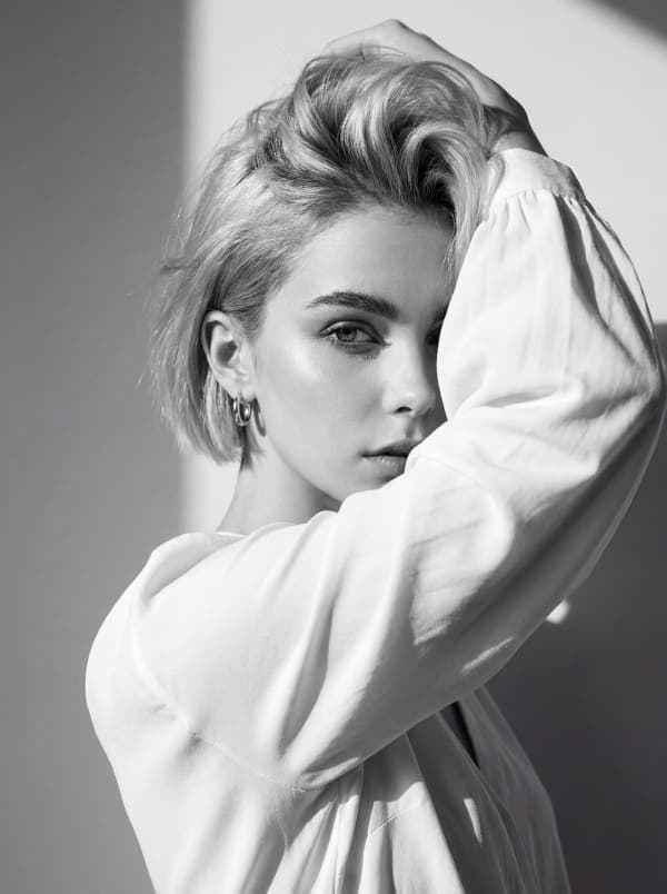 Black and white high-fashion editorial portrait of a woman in a white blouse posing with one arm raised partially covering her face under dramatic sunlight shadows.