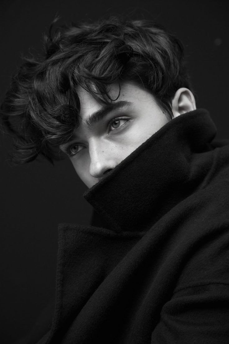 Black-and-white close-up fashion portrait of a young man with tousled dark hair and a raised wool coat collar covering the lower face, photographed against a dark studio background.