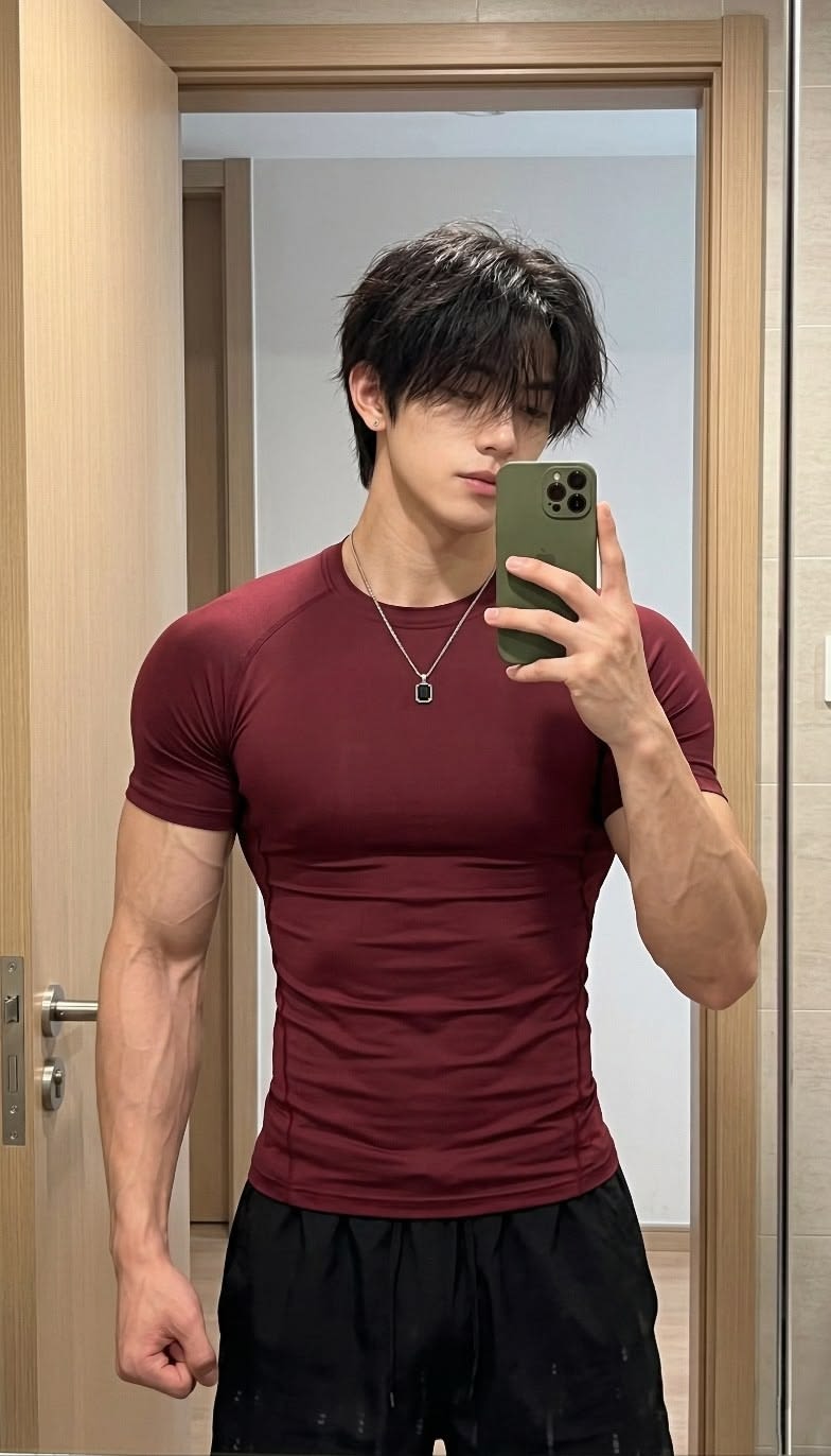 Photorealistic indoor mirror selfie portrait of a young athletic man wearing a fitted deep burgundy compression T-shirt, black athletic shorts, silver pendant necklace, and holding an olive-green smartphone in a clean doorway mirror setting.