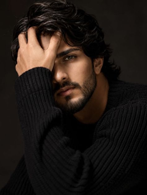 Close-up portrait of a bearded man in a black ribbed sweater with one hand resting across his forehead, lit by soft studio lighting against a dark background.