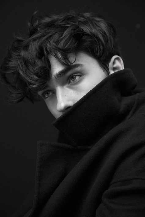 Black-and-white close-up fashion portrait of a young man with tousled dark hair and a raised wool coat collar covering the lower face, photographed against a dark studio background.