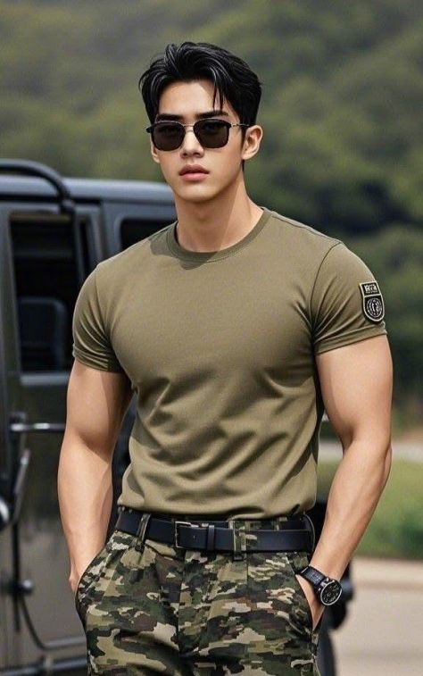 Young man wearing an olive-green military-style t-shirt and camouflage cargo pants, standing confidently beside a roadside with a blurred black SUV and green hills in the background.