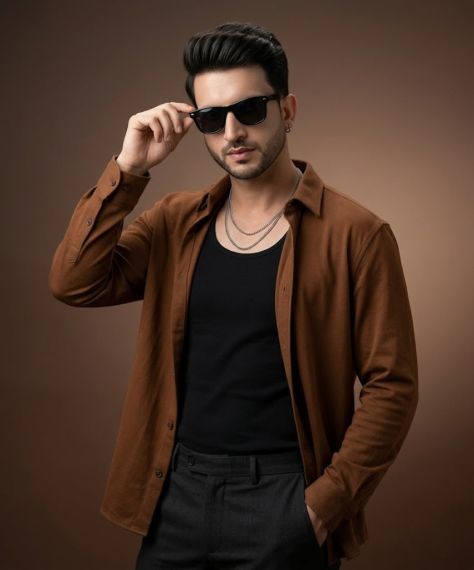 Young South Asian man in his 20s wearing dark sunglasses, chestnut-brown overshirt over a black tank top, holding his sunglasses with one hand and keeping the other hand in his pocket, posed in a warm professional studio with soft gradient lighting.