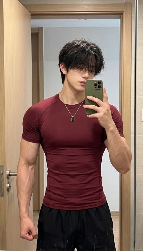 Photorealistic indoor mirror selfie portrait of a young athletic man wearing a fitted deep burgundy compression T-shirt, black athletic shorts, silver pendant necklace, and holding an olive-green smartphone in a clean doorway mirror setting.