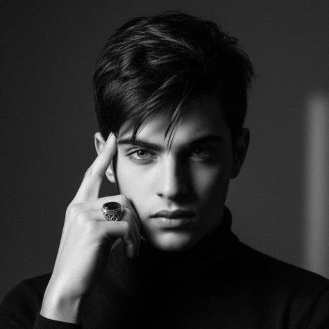 Ultra-photorealistic square black-and-white editorial studio portrait of a young man wearing a black high-neck turtleneck, posing with one hand beside his face and a large dark oval statement ring, with dramatic chiaroscuro lighting and a dark charcoal-gray background.