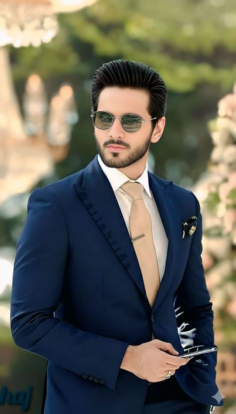 Photorealistic vertical outdoor fashion portrait of a young man wearing a deep navy blue formal suit, white dress shirt, beige satin necktie with tie bar, pocket square, and round metal sunglasses, standing in a softly blurred garden walkway while holding a smartphone near the lower frame.