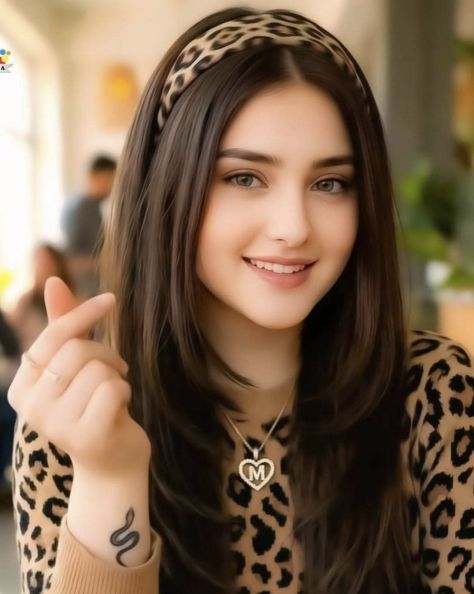 Photorealistic vertical indoor café-style portrait of a young woman wearing a matching leopard-print top and padded headband, smiling at the camera with a finger-heart gesture near her face, two slim rings, a small snake wrist tattoo, and a silver heart pendant necklace with a capital “F”, against a warm softly blurred indoor background.