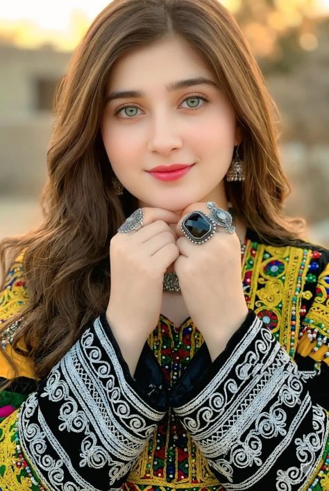 Photorealistic vertical close-up portrait of a young woman wearing a traditional black multicolored Afghan-inspired embroidered dress, dangling silver earrings, and large ornate statement rings, with both hands gently placed under her chin, pale green eyes looking into the camera, and a warm rustic earth-toned outdoor background softly blurred behind her.