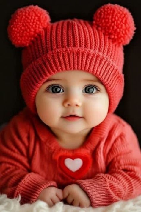 Photorealistic vertical close-up portrait of an infant baby facing the camera, wearing a vivid coral-red knitted winter hat with two fluffy pom-poms and a matching coral-red knitted sweater with a red-and-white heart decoration, with both hands resting together on a soft white fuzzy blanket against a dark studio background.