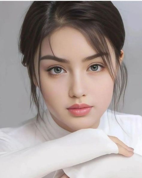 Photorealistic close-up beauty portrait of a young adult woman with blue-gray greenish eyes, dark tied-back hair, glossy rose-pink lips, and a white long-sleeve top, resting her chin on crossed arms against a soft light-gray studio backdrop.