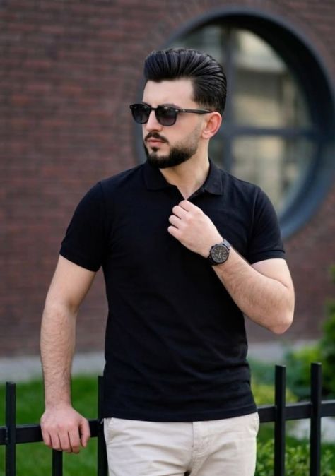 Photorealistic editorial portrait of a man wearing a black polo shirt, beige trousers, and dark sunglasses, standing beside a black metal fence in front of a softly blurred brick building with a circular window, looking confidently off to the side.