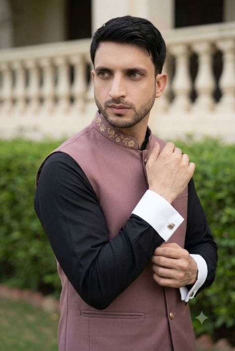 Editorial portrait of a young man wearing a dusty rose embroidered Nehru waistcoat over a black kurta, posing with crossed arms in a landscaped outdoor setting with a softly blurred classical pavilion background.
