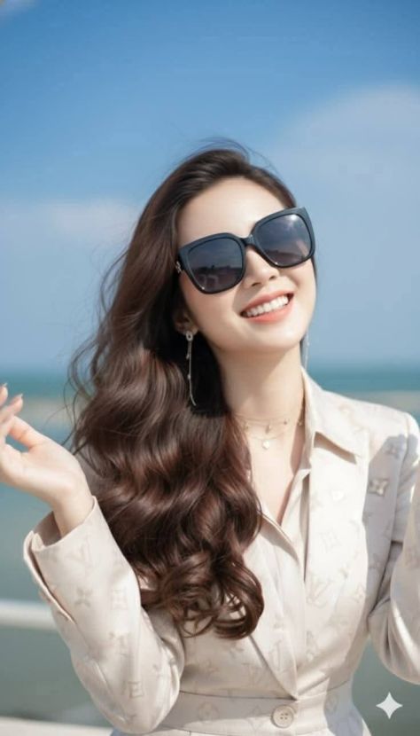 Vertical coastal beauty portrait of a smiling young woman in an ivory belted blazer, oversized dark cat-eye sunglasses, layered necklaces, and long wavy chestnut hair against a softly blurred blue ocean and sky.