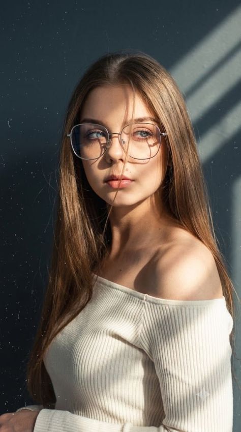 A photorealistic young woman with long dark hair and silver rounded-rectangle glasses wears an ivory off-the-shoulder ribbed top while diagonal window-blind shadows cross her face and exposed shoulder against a dark slate-grey wall.