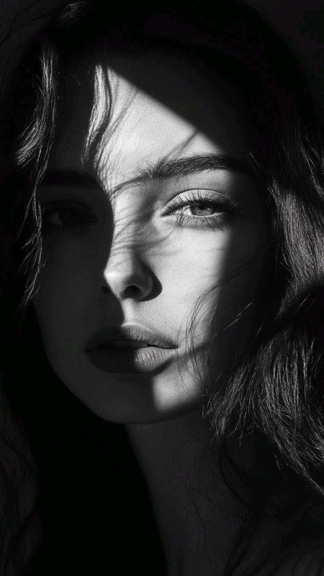 A high-contrast black-and-white close-up portrait of a woman with long dark hair, one eye illuminated by hard directional light and the opposite side of her face fading into a pure-black background.