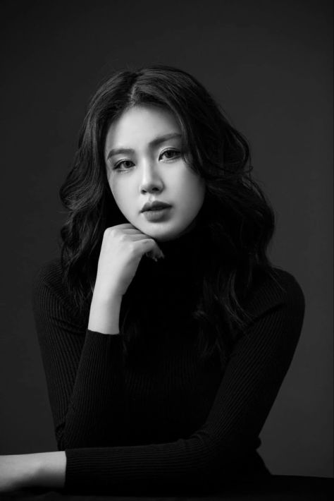 A photorealistic black-and-white studio portrait of a woman in a fitted ribbed turtleneck resting her chin on one curled hand while looking directly into the camera.