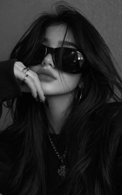 A high-contrast black-and-white editorial portrait of a feminine-presenting young adult wearing oversized opaque sunglasses, a charcoal knit top, metallic jewelry, and long pointed black nails while resting her chin against one hand.