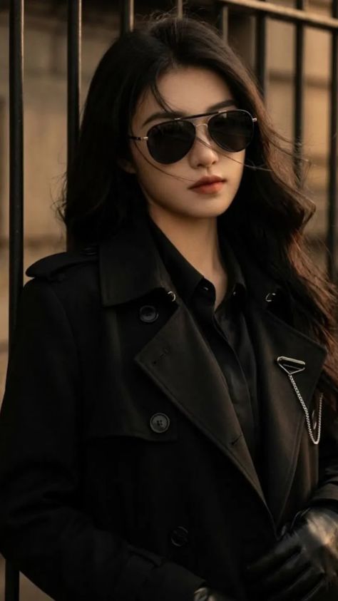 A photorealistic feminine-presenting young adult in an all-black double-breasted trench coat and aviator sunglasses leans against an iron railing in a warmly graded urban setting.