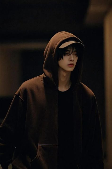 A photorealistic masculine-presenting young adult with blue-black wispy hair and light facial stubble wears an oversized espresso-brown hoodie over a dark cap in a dim, warmly lit architectural interior.
