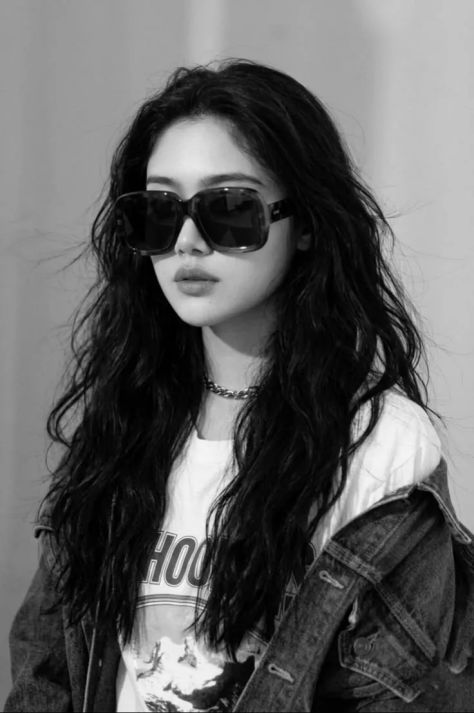 A black-and-white fashion editorial portrait of the same young woman from the uploaded reference wearing oversized dark sunglasses, a dropped dark denim jacket over a white graphic T-shirt, a chunky curb-chain necklace, and voluminous wavy hair against a pale gray studio background.