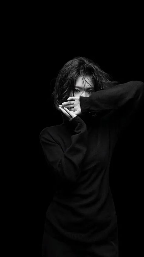 A high-contrast black-and-white minimalist editorial portrait of the same young woman from the uploaded reference with narrowed direct gaze, tousled dark hair, crossed hands concealing the lower face, an oversized black knit sweater, and a seamless black studio background.
```